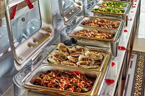Các khay hâm nóng trên buffet du thuyền Vịnh Hạ Long bày hàu nướng, hải sản xào và rau củ.