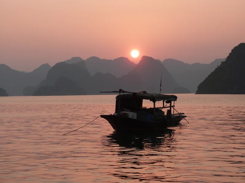 Một chiếc thuyền gỗ nhỏ trôi trên mặt nước tĩnh lặng khi hoàng hôn buông xuống sau những ngọn núi đá vôi ở vịnh Hạ Long